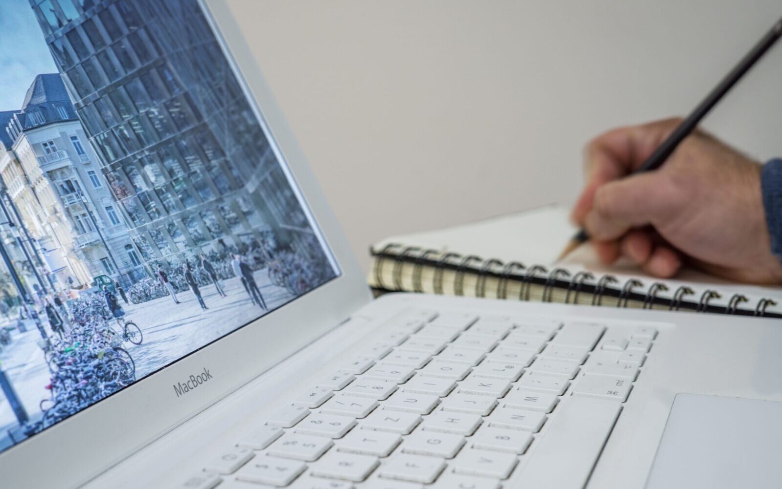 A person taking notes in a notebook beside an open laptop displaying an urban scene.