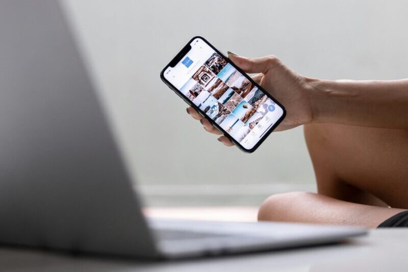 A woman multitasks using a smartphone and laptop for social media in a modern setting.